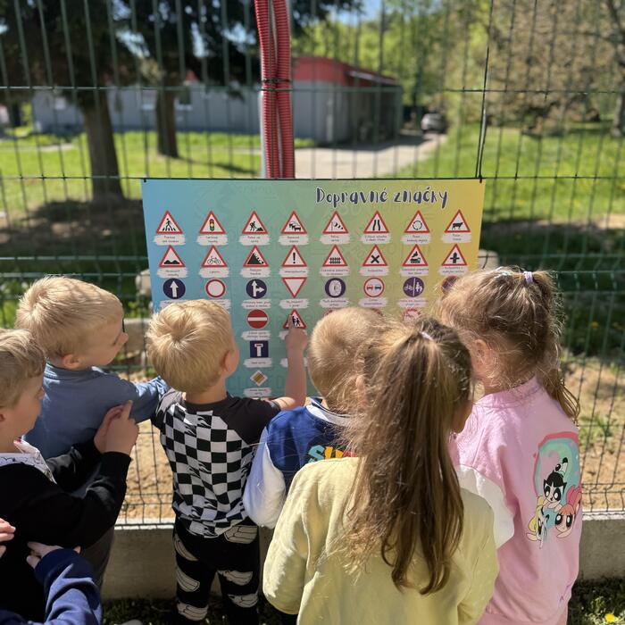 Deti z materskej školy pri plote ukazujú na edukačnú tabuľu Dopravné značky