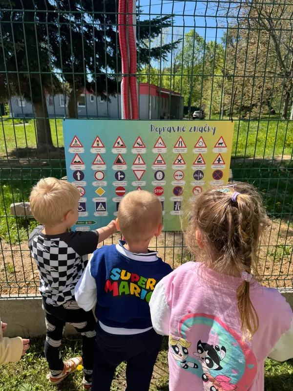 Deti v materskej škole si pri plote prezerajú tabuľu s dopravnými značkami